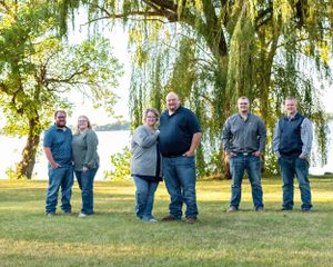 Family of six posing casually on a sunny lawn with a weeping willow and lake backdrop.