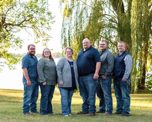 Six adults in coordinated casual outfits smile together under a golden weeping willow tree by a lake.