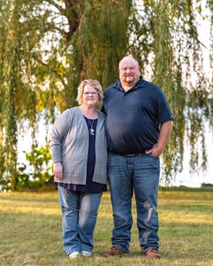 Smiling couple standing together outdoors under a weeping willow tree by a lake.