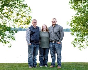 Three siblings smiling together outdoors by a lakeside with trees in background.