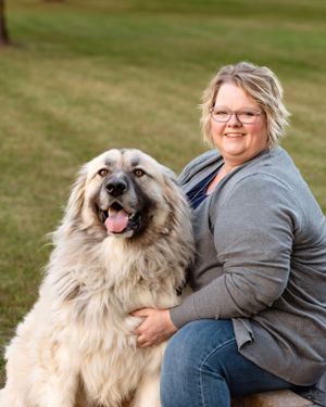 Smiling woman sitting outdoors on grass with a large fluffy Great Pyrenees dog.