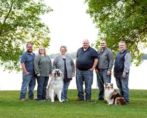 Family group of six people with two dogs posing outdoors near a lakeside.
