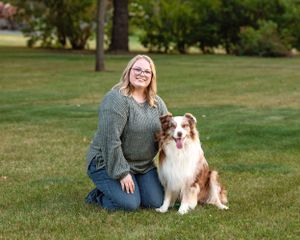 Young woman kneeling on grass smiling with a brown and white Australian Shepherd dog.
