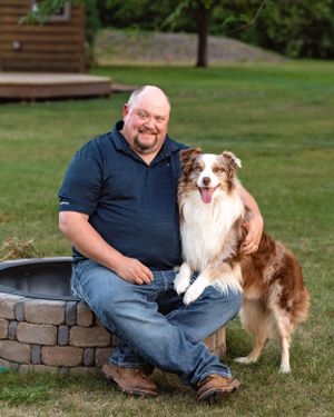 Smiling man sitting by a fire pit outdoors with a brown Australian Shepherd dog.
