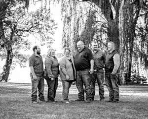 Black and white photo of six adults laughing together under large trees near a lakeside setting.