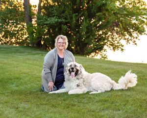 Woman sitting on grass smiling outdoors with a fluffy white dog near a lake.