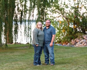 Couple smiling together on green lawn under weeping willow tree near lake on sunny day.