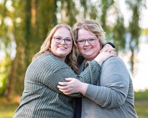 Two women wearing glasses hugging and smiling outdoors in a park setting.