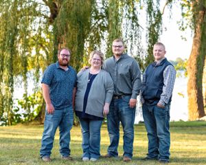 Family of four smiling outdoors near a willow tree by a lake on a sunny day.