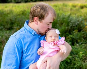 Father in blue shirt tenderly kissing his newborn baby dressed in pink in an outdoor field setting.