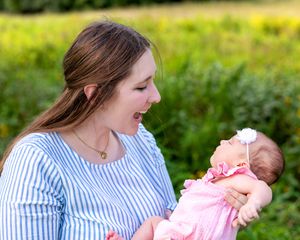 Young mother smiling lovingly at her newborn baby in pink dress while standing in a sunny outdoor field.