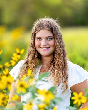 Smiling teenage girl with curly hair posing among vibrant yellow wildflowers in a bright outdoor setting.