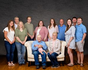 Multi-generational family portrait with grandparents seated on a loveseat holding a newborn baby, surrounded by relatives.