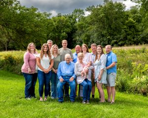 Extended family group photo outdoors on green grass with lush trees, two elders seated center holding a newborn in pink.