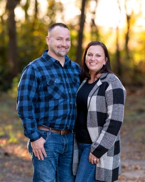 Smiling couple in plaid shirts posing outdoors among autumn trees at golden hour.