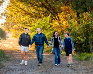 Family of four walking hand-in-hand down a scenic dirt path lined with fall foliage.