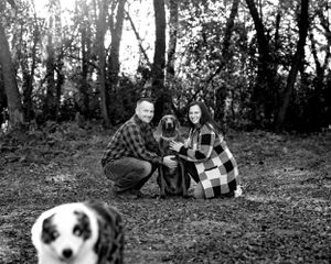 Black and white photo of couple crouching with their dogs in a wooded outdoor setting.
