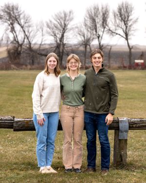 Three young adult siblings smiling together leaning on a rustic fence on a farm.
