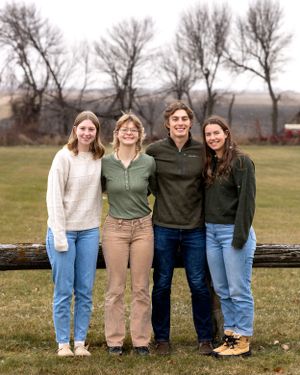 Four young adult siblings smiling together beside a rustic wooden fence on a farm.