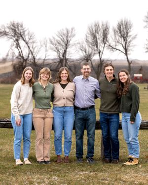 Family of six smiling together in a scenic farm setting with bare winter trees behind them.
