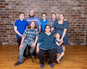 Large family group portrait with eight members posing in front of a rustic brick wall on a wood floor.