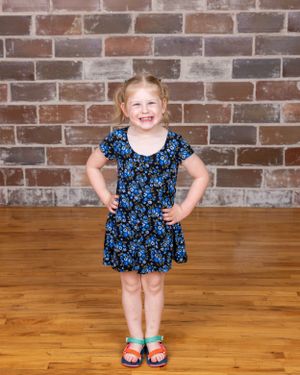 Smiling young blonde girl in a blue floral dress posing with hands on hips against a brick wall.