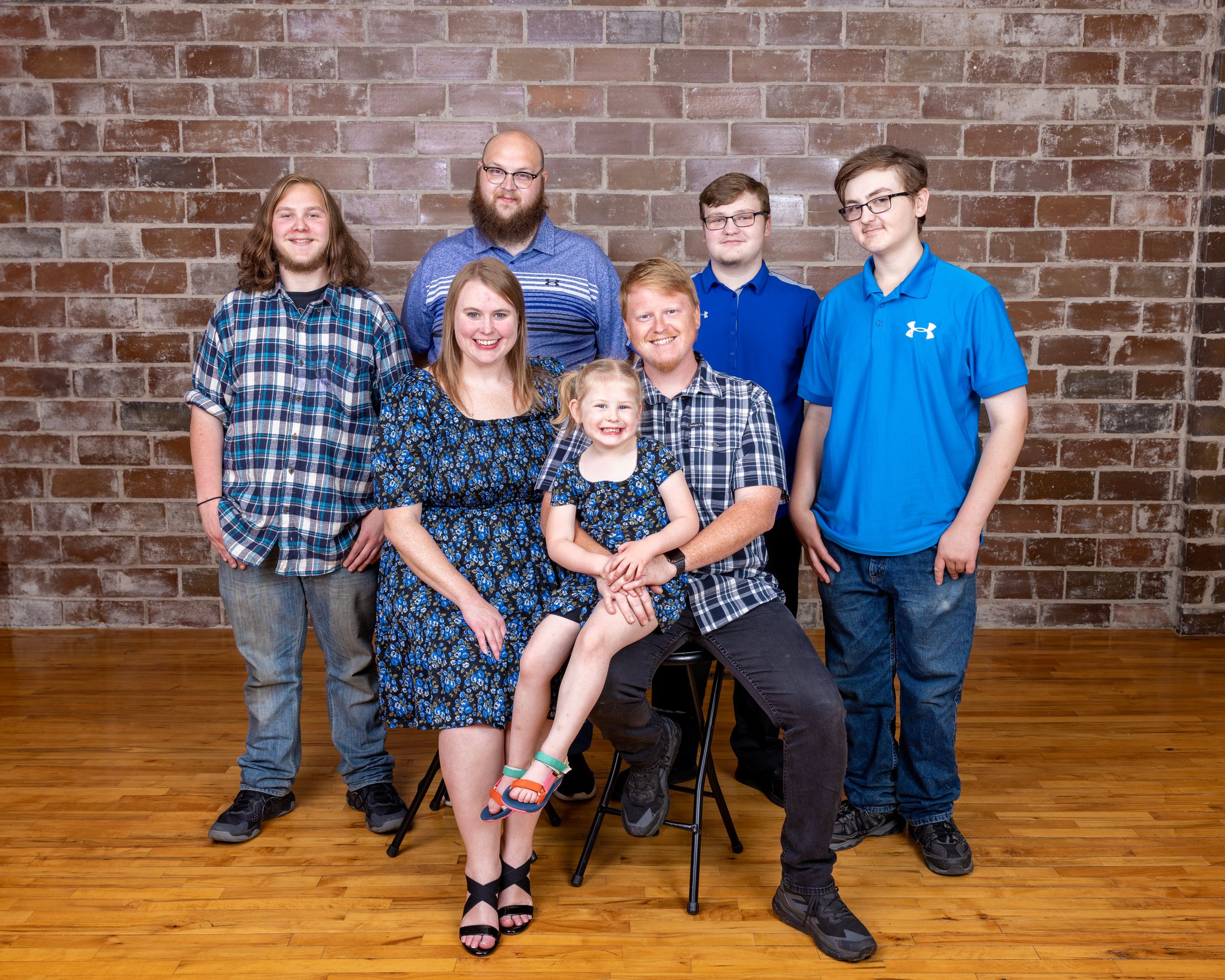 Extended family group of seven posing together in front of a rustic brick wall on hardwood floors.
