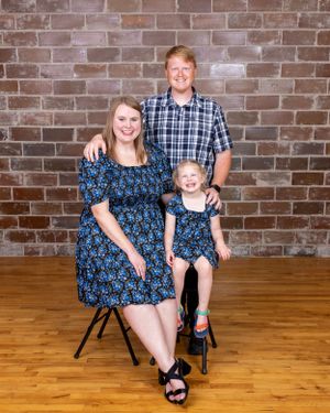 Family portrait of parents and young daughter in matching blue floral dresses seated before a brick wall.