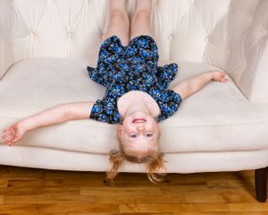 Playful young girl in a blue floral dress lying upside down on a white tufted sofa laughing.