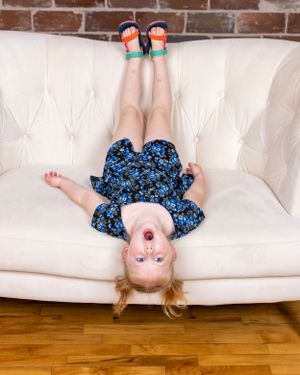 Young girl in a blue floral dress lying upside down on a white tufted sofa with feet in the air.