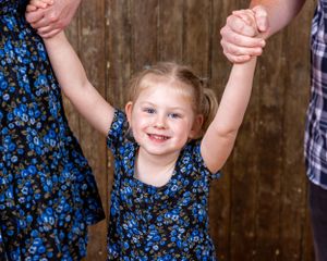 A happy little girl in a floral dress holds both hands up while standing between two adults.