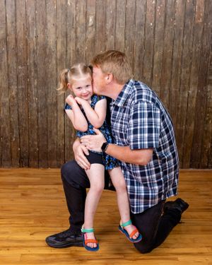 A man kneels to kiss a smiling little girl in a floral dress on the cheek.