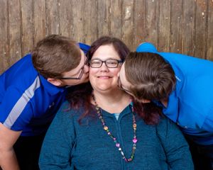 Two young men in blue shirts kiss a smiling woman on the cheeks in a sweet portrait.