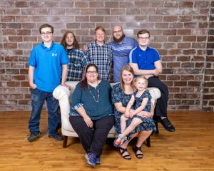 A large family of eight poses together in front of a brick wall for a group portrait.