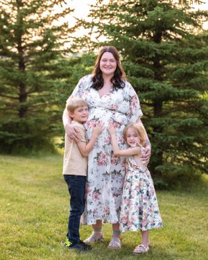 Pregnant woman in floral dress with two children hugging her outdoors at sunset.