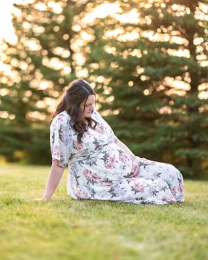 A pregnant woman in a white floral dress sits on green grass, gazing at her baby bump at golden hour.