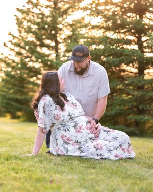 A pregnant woman reclines on grass in a floral dress as her partner gazes at her lovingly at sunset.