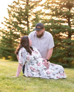 A couple shares an intimate moment on the grass, the pregnant woman in a floral dress leaning back at sunset.