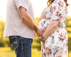 A tattooed couple holds hands while the pregnant woman cradles her baby bump in a floral dress at golden hour.