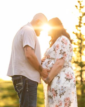 A couple holds hands facing each other with a glowing sunset behind them during a maternity photo session.
