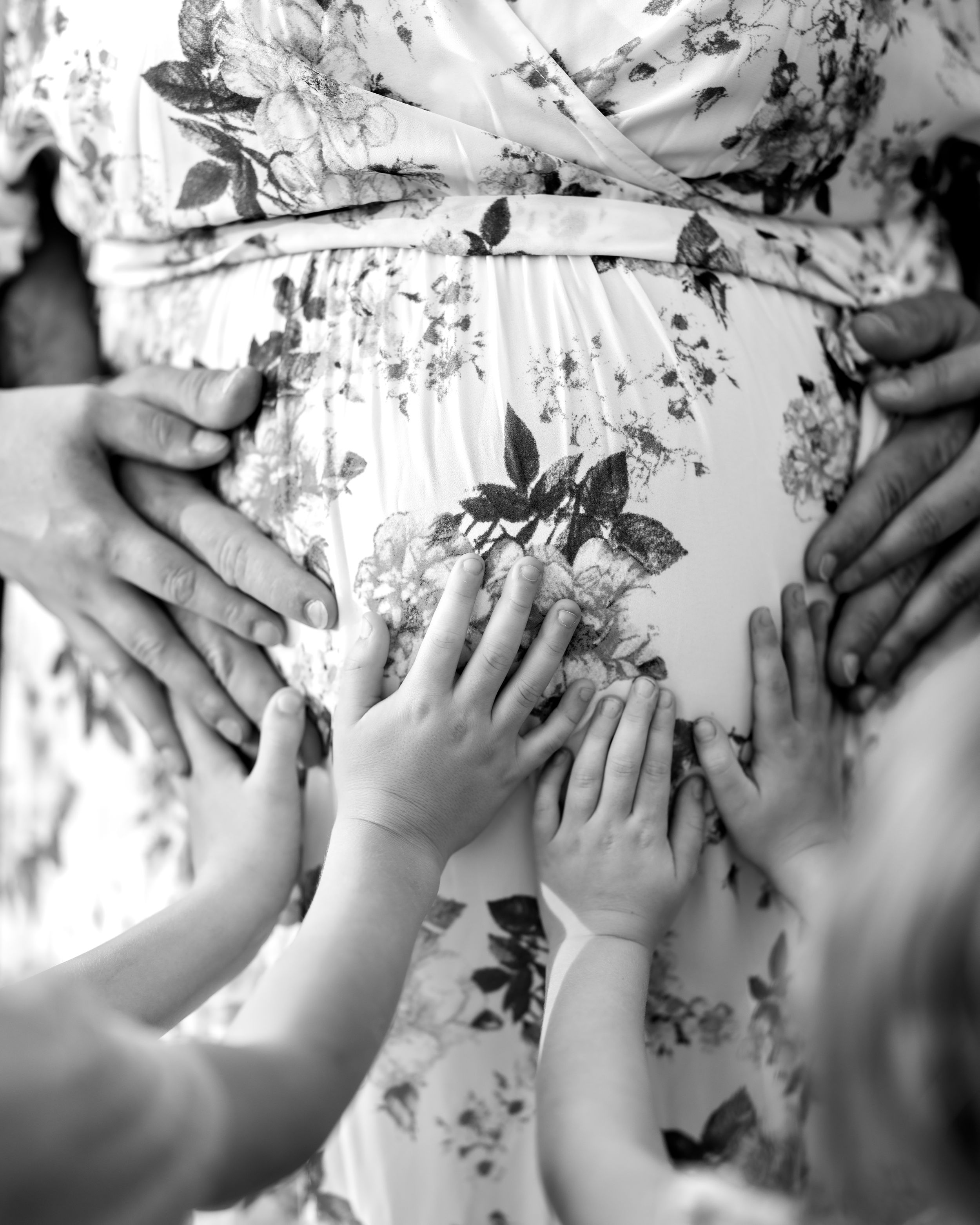 Black and white close-up of multiple hands, including small childrens hands, touching a pregnant belly in a floral dress.