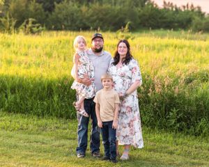 Family maternity photo session in golden hour field, pregnant mother in floral dress with husband and two children.