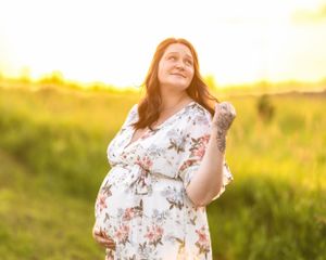 Glowing pregnant woman in floral dress looking up joyfully during sunny outdoor maternity photo session.