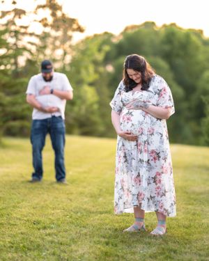 Pregnant woman in floral dress with man standing behind her in sunny green field.
