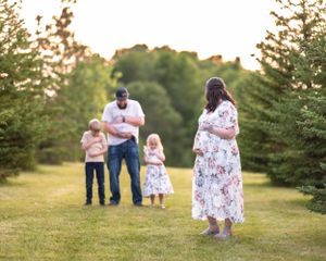 Family with pregnant mother and two children standing in tree-lined field at sunset.