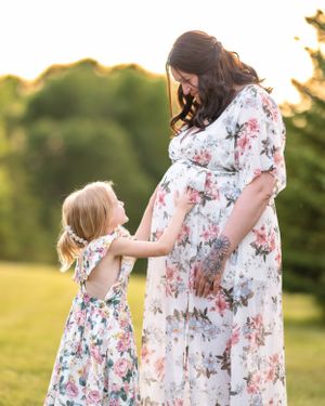 Little girl in floral dress touching pregnant mother's belly in golden sunset light.