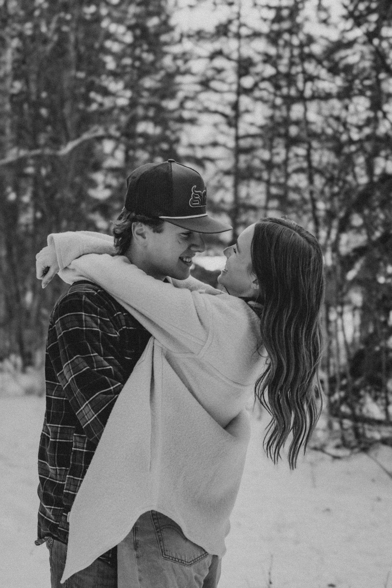 Black and white photo of a couple hugging and smiling in a snowy winter forest.