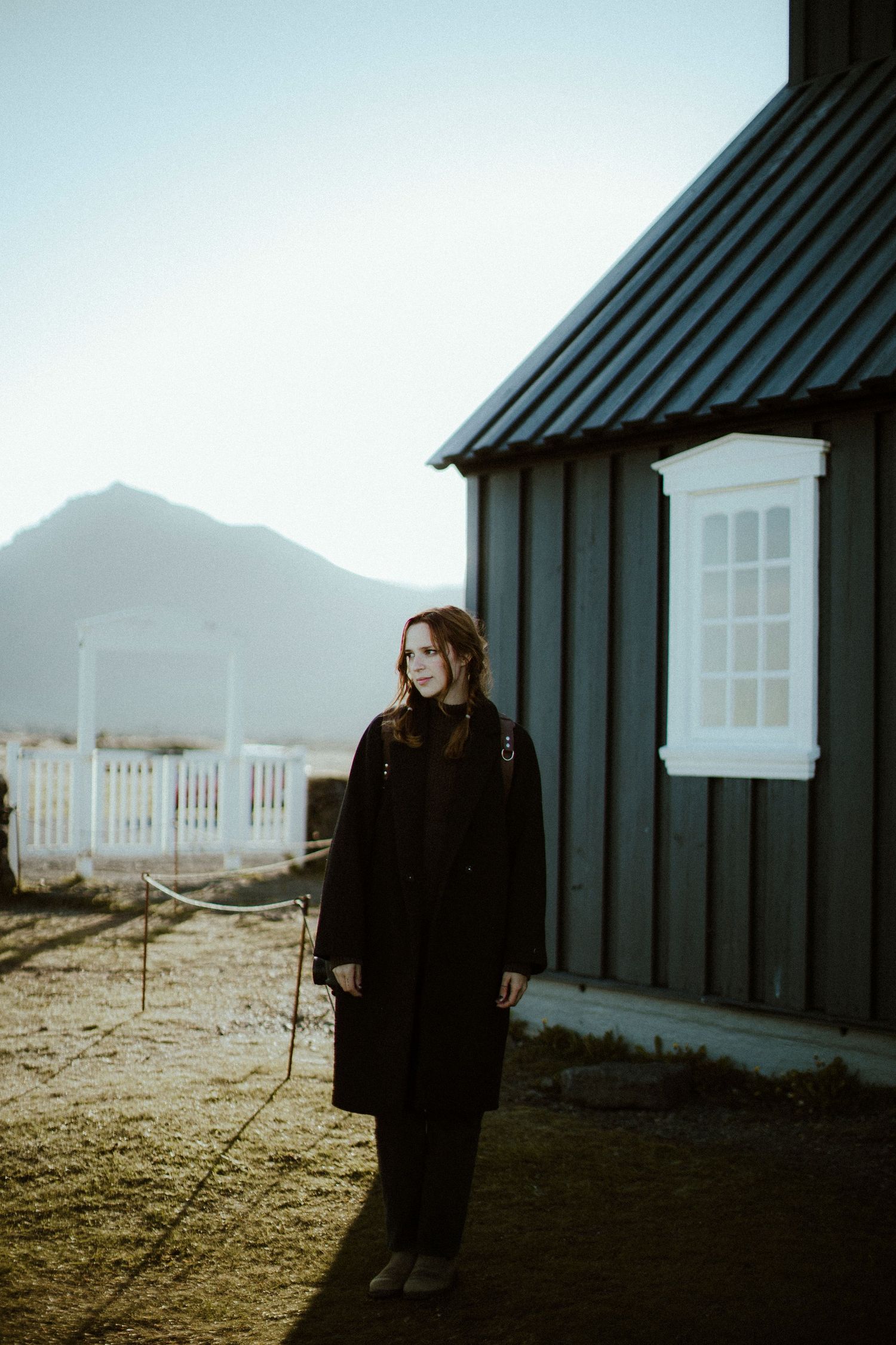 Destination elopement photographer in black coat stands beside rustic building with mountains in background
