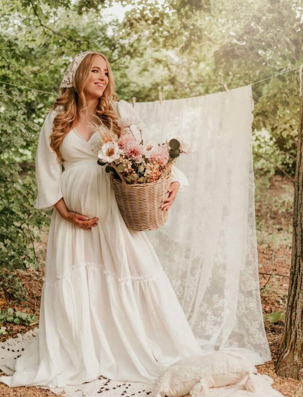 Ethereal maternity session in white flowing dress with flower basket in outdoor garden setting.