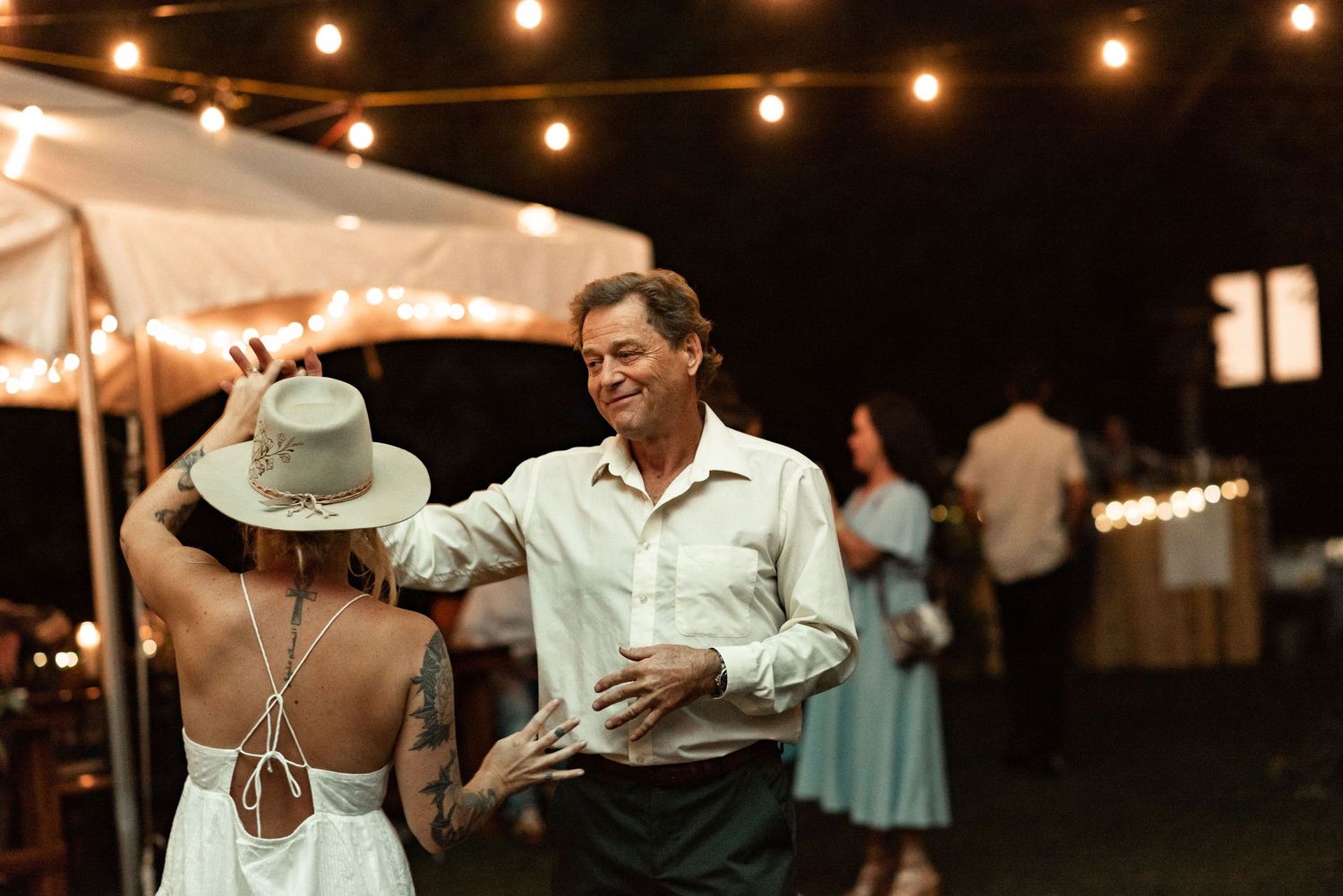 Evening wedding reception scene with dancing couple under string lights and tent.
